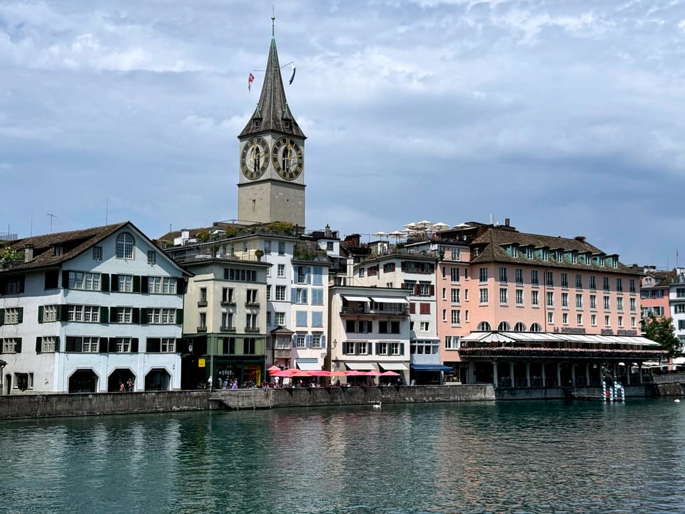 Calm waterfront scene in Zurich’s old town—St. Peter’s church clock tower rises above pastel-colored historic buildings and café terraces lining the Limmat River, with their reflections